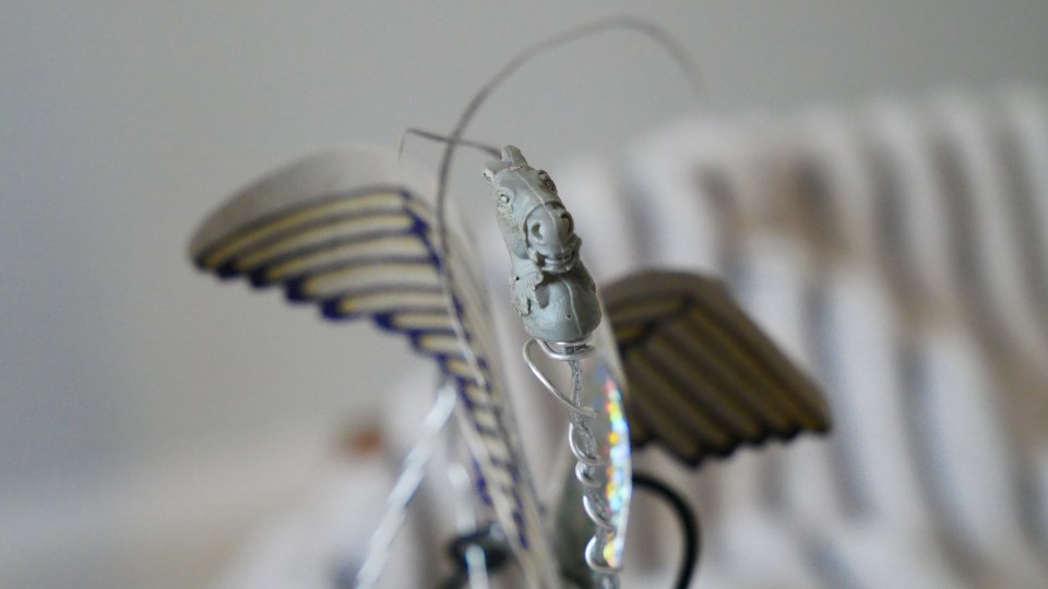 A small, old grey plastic toy horse head faces forward, with hand-drawn wings and a wireframe body
